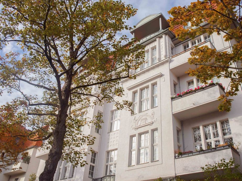 White residential building with balconies, surrounded by trees with autumn leaves, under a partly cloudy sky.