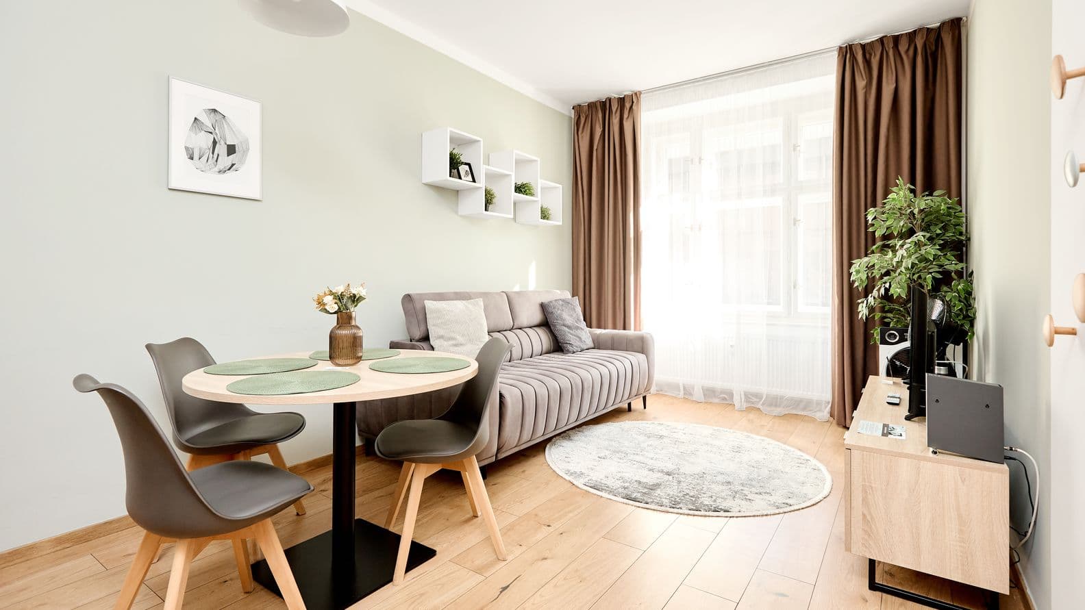Modern living room with wooden flooring. A round dining table with three grey chairs and a vase of flowers stands by the left wall. A grey sofa with cushions is placed along the wall beneath white floating shelves. Opposite, there is a low TV cabinet with a television and a tall indoor plant beside it. The space is filled with natural light from a large window with sheer curtains and brown drapes.