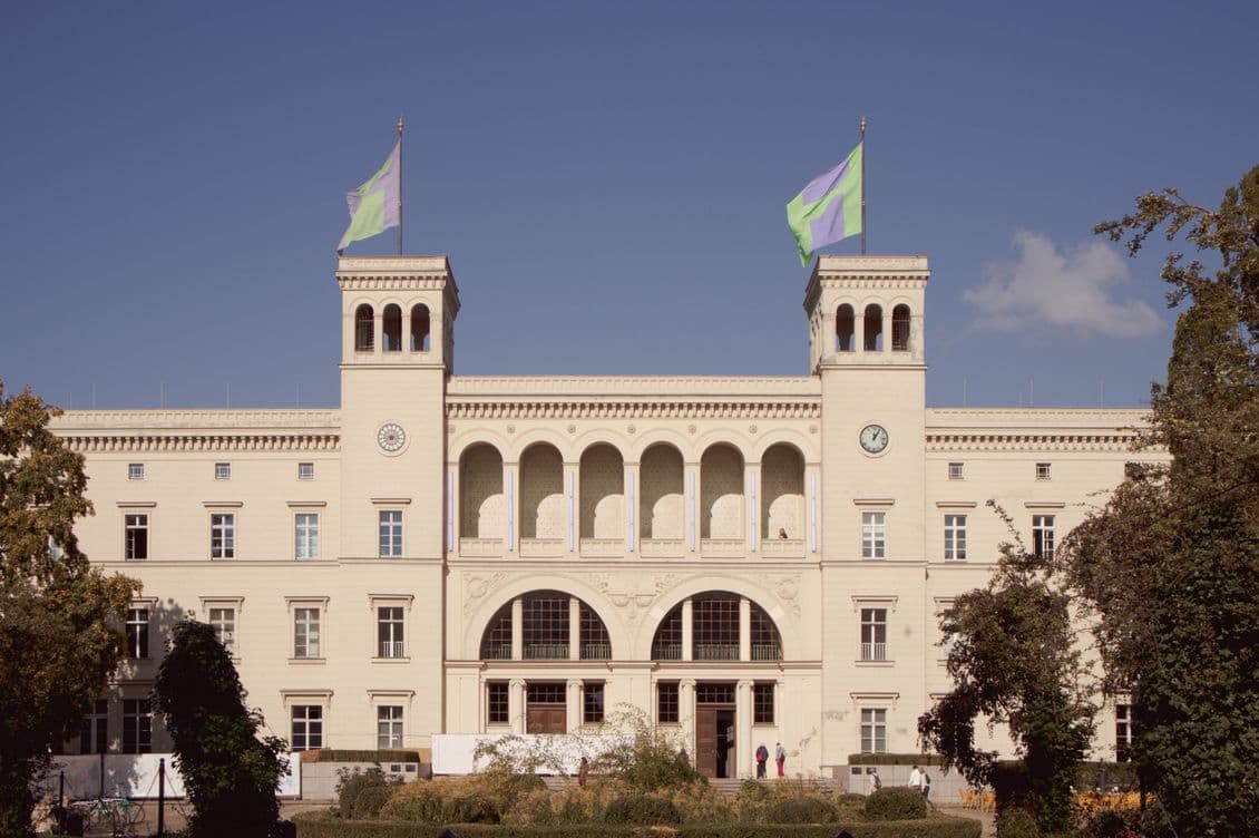Historic building with two towers, arched windows, and two flags on top, set against a clear blue sky with trees in the foreground.