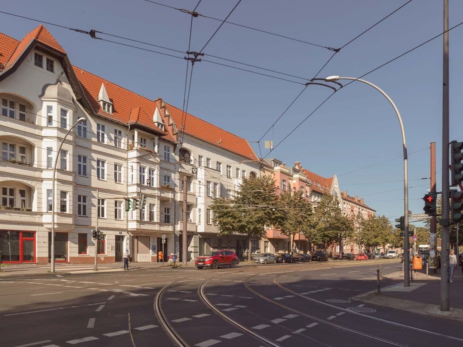 Street scene with historic buildings, red roofs, tram tracks, and traffic lights under a clear blue sky. Trees line the sidewalk.