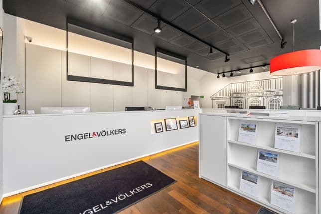 Modernes Immobilienbüro mit weißen Schreibtischen, Wandkunst und einer roten Deckenleuchte. „Engel & Völkers“-Logo an prominenter Stelle.