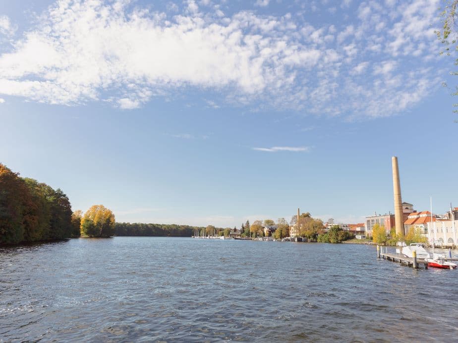 A serene river scene with boats docked, surrounded by trees and buildings under a partly cloudy blue sky.