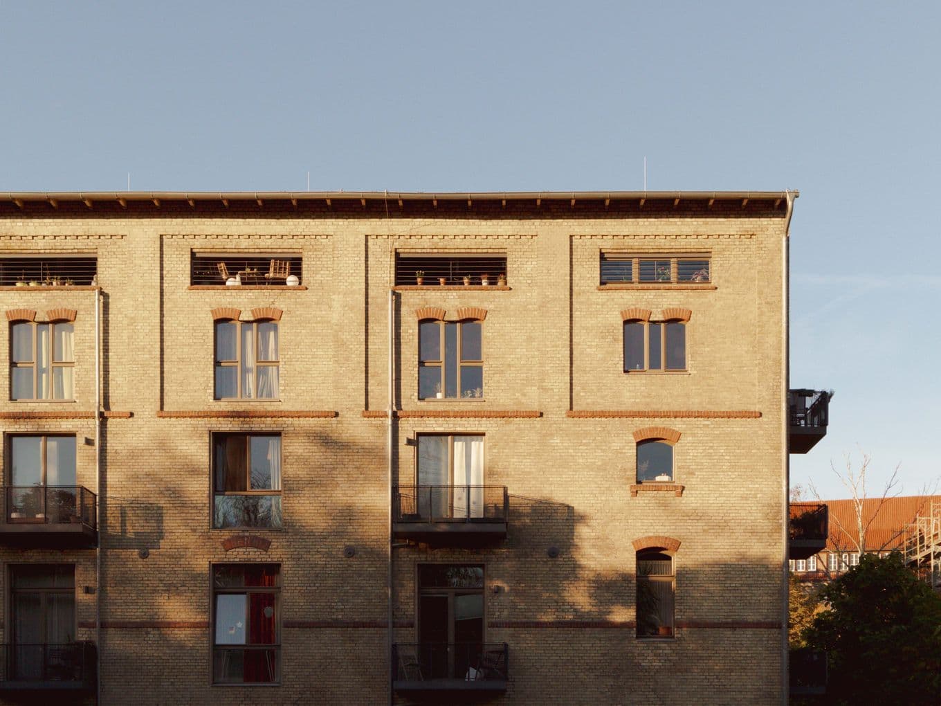 Brick apartment building with balconies and large windows, bathed in warm sunlight. Shadows of trees are visible on the facade.