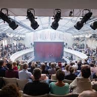 Vollbesetzter Theatersaal mit historischer Architektur, Bühnenbild mit rotem Vorhang und professioneller Beleuchtungstechnik