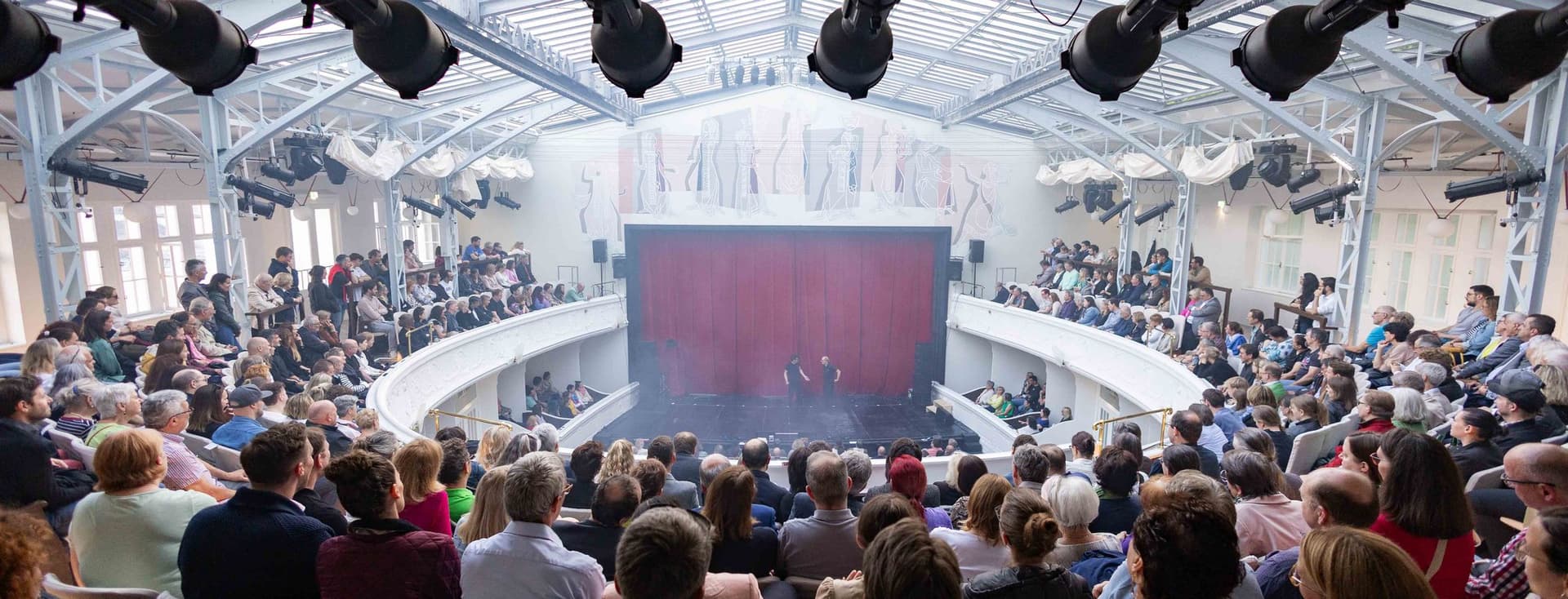 Vollbesetzter Theatersaal mit historischer Architektur, Bühnenbild mit rotem Vorhang und professioneller Beleuchtungstechnik