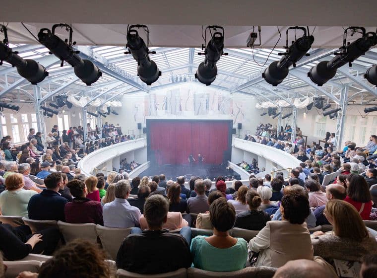 Vollbesetzter Theatersaal mit historischer Architektur, Bühnenbild mit rotem Vorhang und professioneller Beleuchtungstechnik