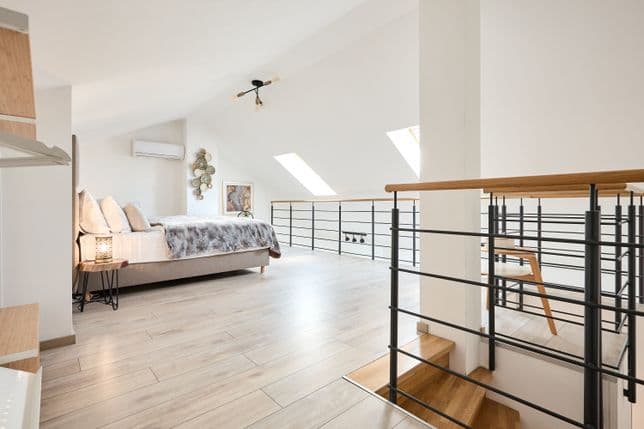 Bright attic bedroom located in an open gallery above a staircase. On the left there is a double bed with an upholstered frame, light bedding and a decorative throw. A small bedside table with a lamp stands next to the bed. Two roof windows set into the sloped ceiling bring natural light into the space. A wall-mounted air conditioning unit and decorative elements are visible on the wall. On the right, a railing with a wooden handrail overlooks the stairs leading to the lower floor. The flooring has a light wood finish.