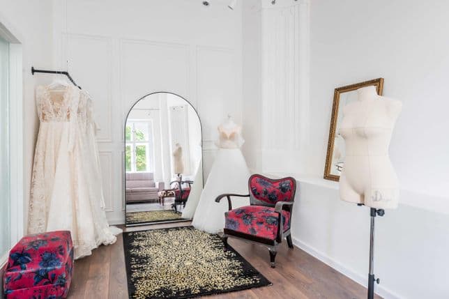 Bridal boutique with white dresses on hangers and mannequins, a black and gold rug, red floral armchairs, and a large mirror. The room is bright with white walls and wooden flooring.