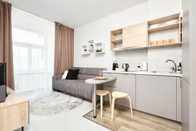 Bright living area connected to a kitchenette. Along the right wall there is a grey kitchen unit with a sink, hob and upper cabinets in light wood finish, with open shelves displaying cups and glasses. A narrow breakfast bar with two light wooden stools stands in front of the kitchen. Near the window with sheer white curtains and brown drapes is a grey sofa with cushions, placed next to a round rug. White wall shelves with small plants hang above the sofa.
