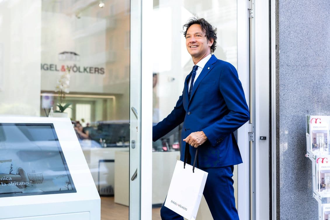 Gianluca Torre apre la porta dell’agenzia Engel & Völkers indossando un abito blu e tenendo in mano una busta con il logo E&V.