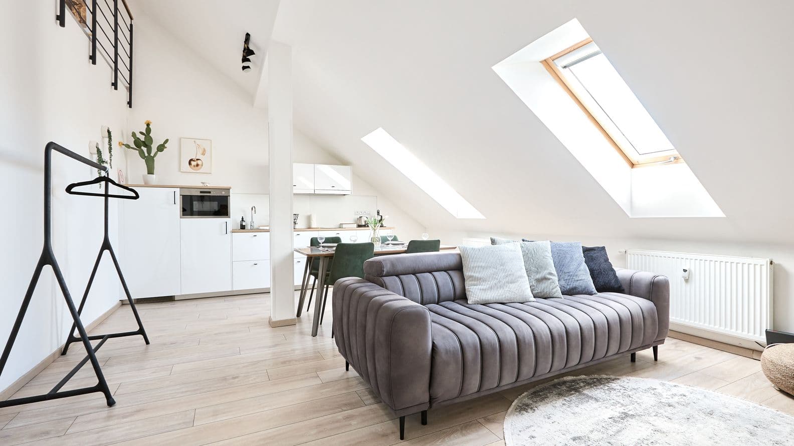 Bright attic interior with a sloped ceiling and two roof windows. In the foreground there is a grey sofa with several cushions, placed next to a round rug on light wood flooring. At the back of the room there is a white kitchenette with upper cabinets and a dining table with green chairs. A radiator runs along the wall, and a simple clothes rack stands nearby. The space feels airy and is filled with natural daylight.