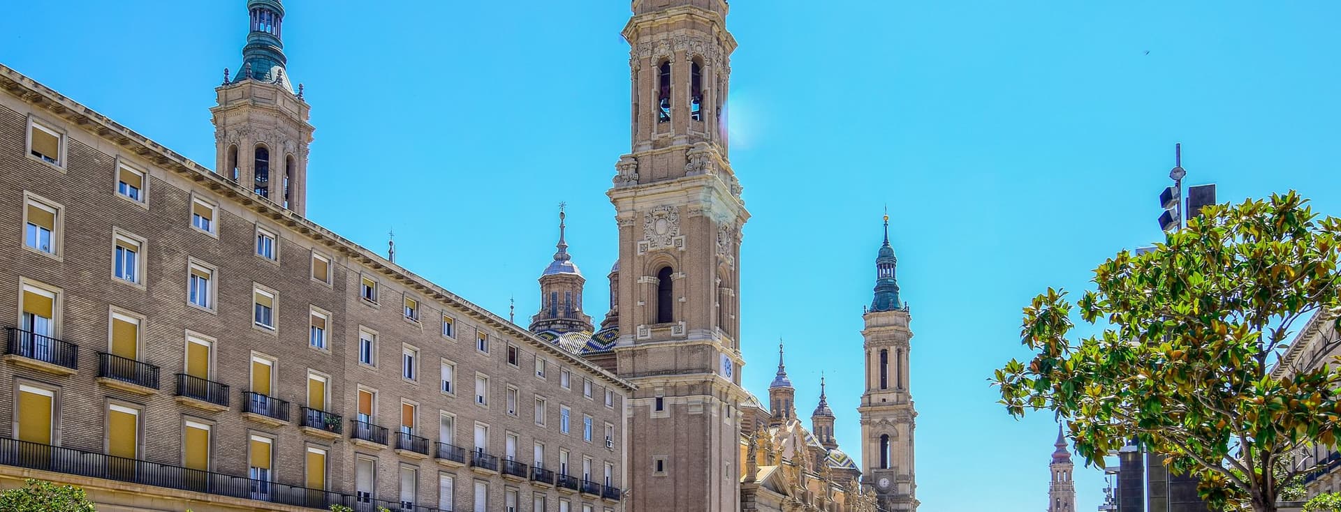 Basilica de Nuestra Señora del Pilar in Zaragoza, Spain, with towers, domes, and a clear blue sky. Trees and a plaza with people are in the foreground.