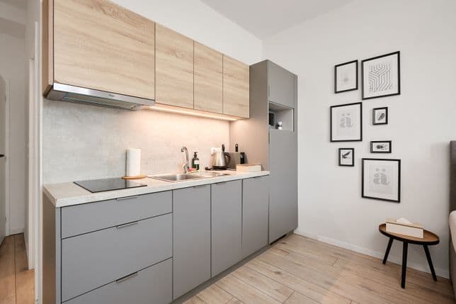 Modern kitchenette with grey lower cabinets and upper cabinets in a light wood finish. The countertop includes a sink with a tap, a hob, an electric kettle and a soap dispenser. Integrated lighting is installed beneath the upper cabinets. On the right there is a tall cabinet with an open shelf. A set of black and white framed pictures hangs on the adjacent white wall, with a small round side table placed below. The floor has a light wood finish.