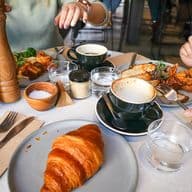A table with two people enjoying brunch, featuring a croissant, coffee, and a dish with meat and greens. One person is using a pepper grinder.