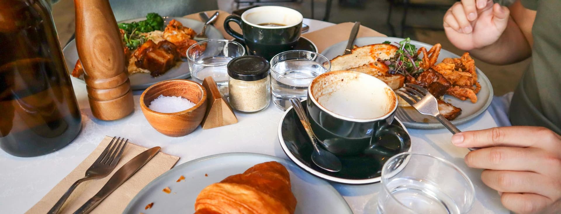 Een tafel met twee mensen die genieten van een brunch, met een croissant, koffie en een gerecht met vlees en groenten. Eén persoon gebruikt een pepermolen.