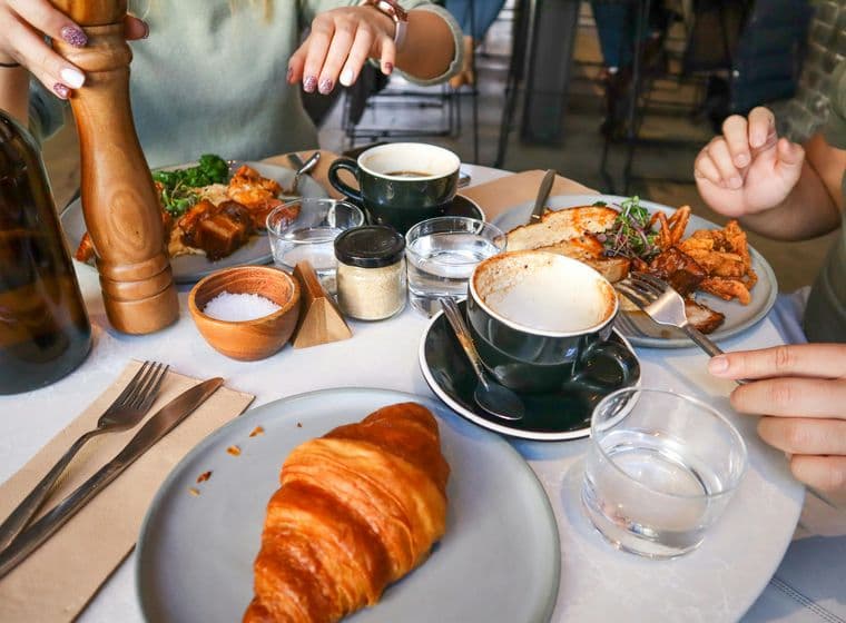 A table with two people enjoying brunch, featuring a croissant, coffee, and a dish with meat and greens. One person is using a pepper grinder.