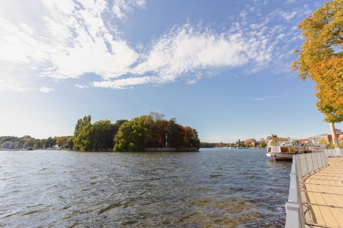 A serene riverside view with a small, tree-covered island under a blue sky with scattered clouds. A boat is docked near the walkway.