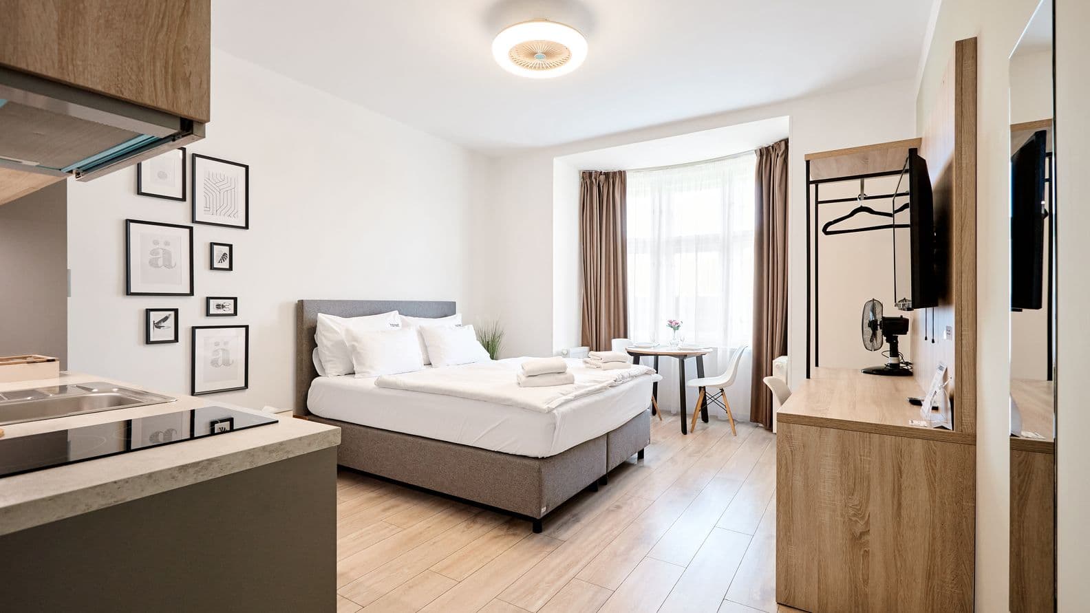 Bright studio interior with wooden flooring. In the centre of the room there is a double bed with an upholstered headboard and white bedding, with folded towels placed on top. On the left, part of the kitchenette with a countertop and hob is visible. Several black and white framed pictures hang on the wall above the bed. By the window with sheer white curtains and brown drapes stands a small round dining table with two chairs. On the right side there is a wooden dresser with a television, a wall mirror and an open clothes rack.