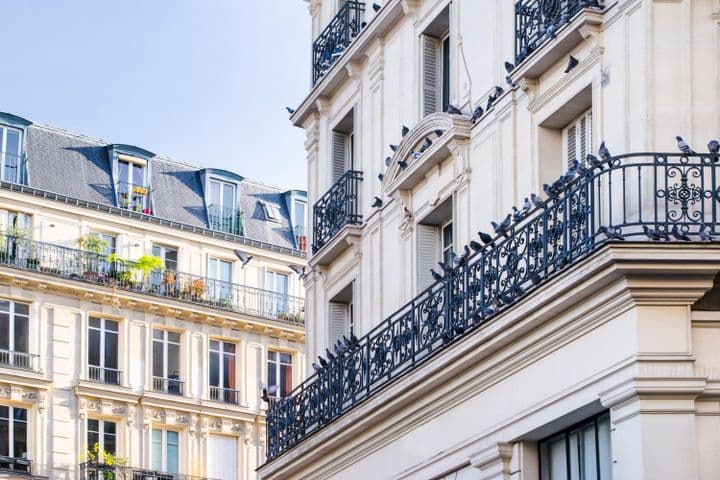 Exterior view of two cream-colored buildings with black wrought iron balconies and pigeons perched on the railings.