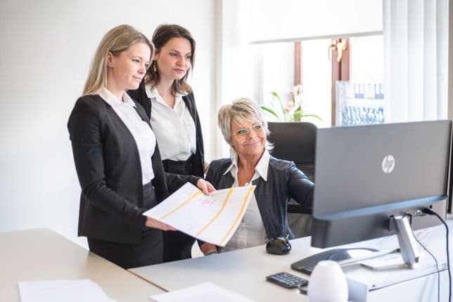 Three professionally dressed women looking at a computer screen in the Engel & Völkers office. One is holding a printed table with color-coded data. The scene highlights teamwork and a detailed analytical approach.