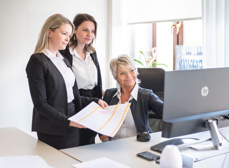 Three professionally dressed women looking at a computer screen in the Engel & Völkers office. One is holding a printed table with color-coded data. The scene highlights teamwork and a detailed analytical approach.