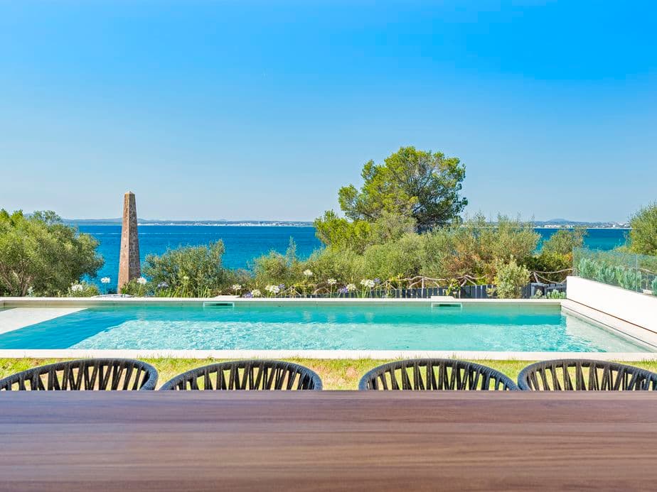 View from a villa in Puerto Alcudia featuring an outdoor dining table, a swimming pool, lush greenery, and the Mediterranean Sea in the background under a clear blue sky.