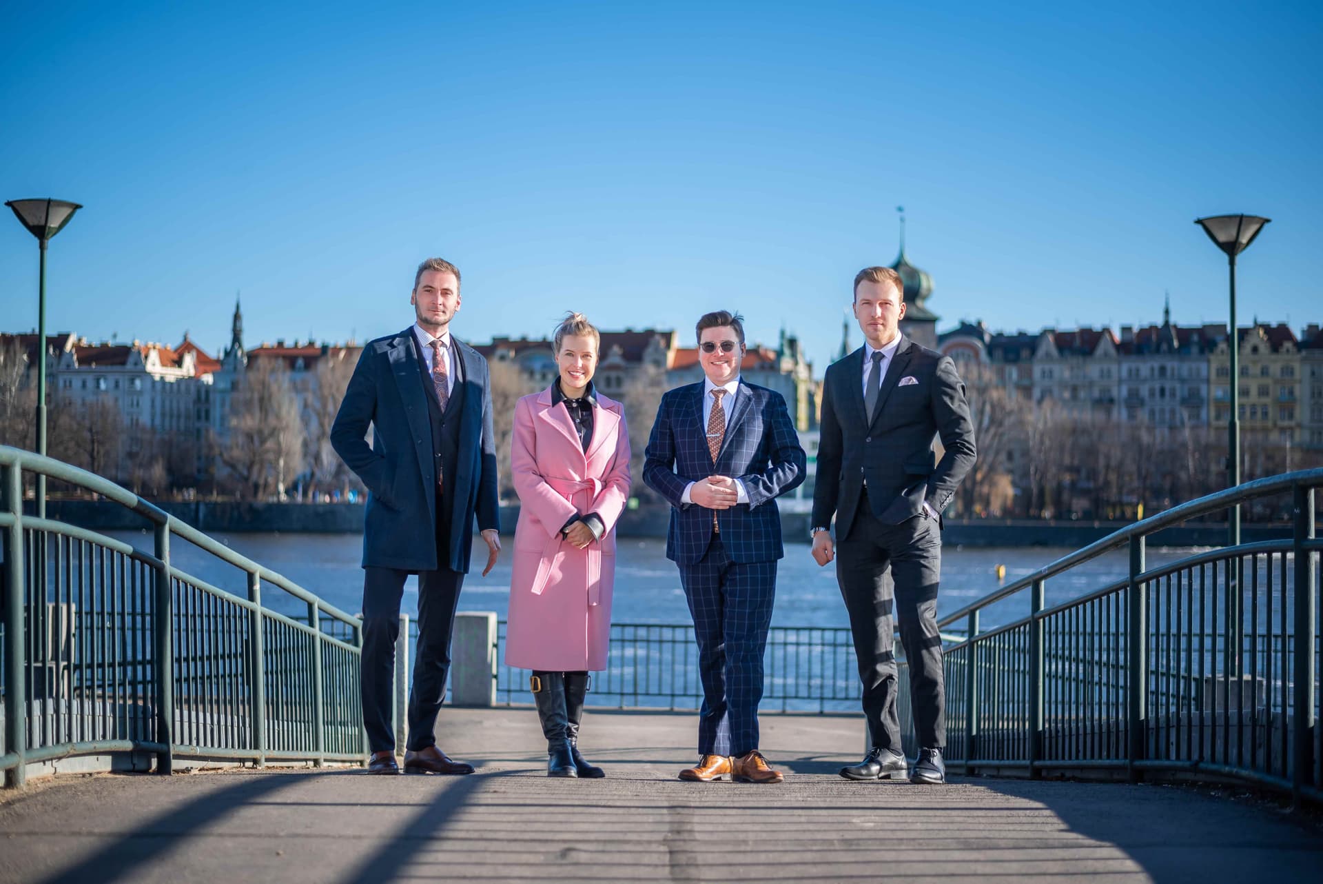 Skupina čtyř realitních specialistů stojí na nábřeží s historickými budovami Prahy v pozadí. Dva muži v oblecích, jedna žena ve světle růžovém kabátu a jeden muž v kostkovaném obleku. Jasná modrá obloha.
