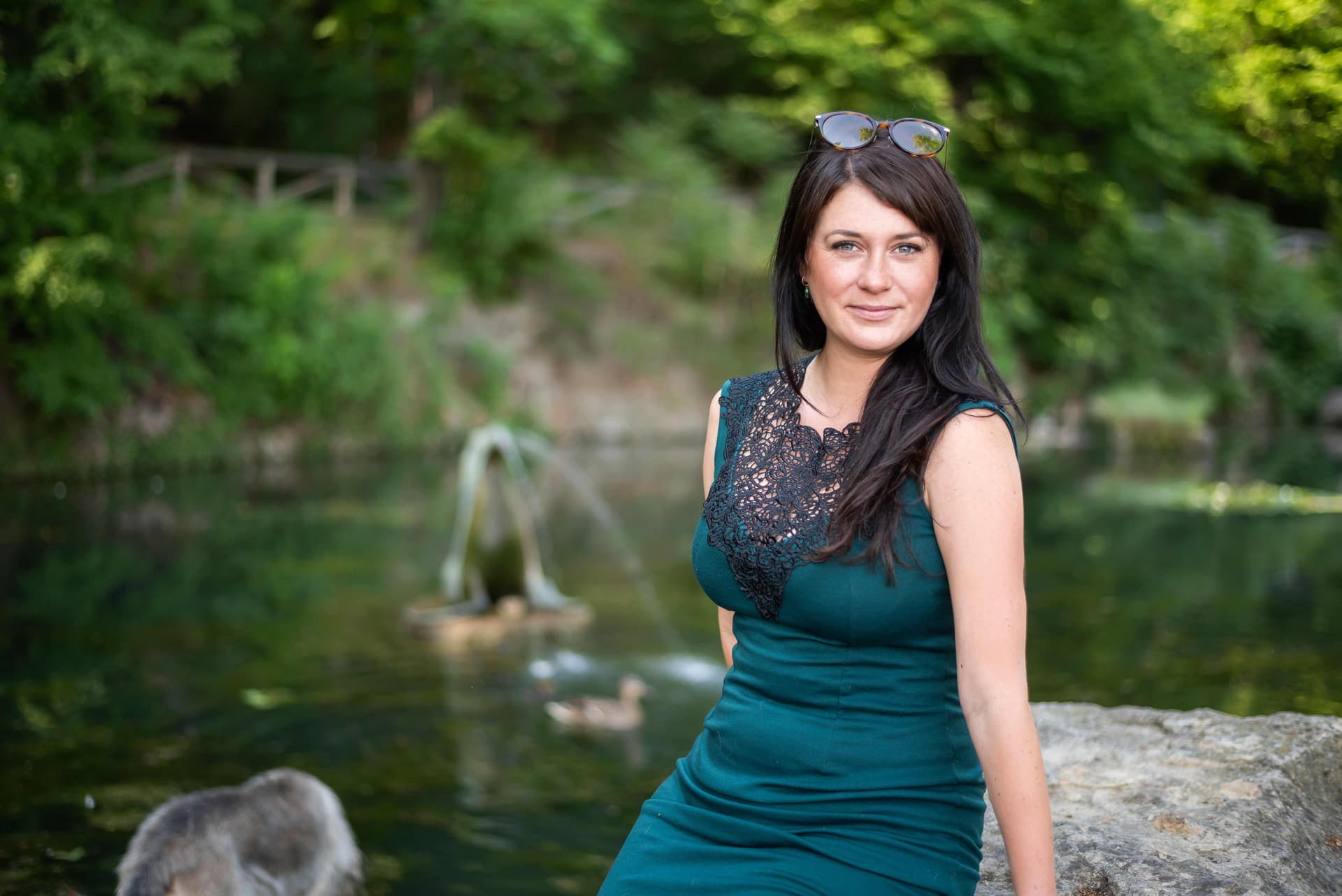 Woman in a dark green dress sitting by a pond with a fountain and ducks. Trees and natural surroundings in the background.