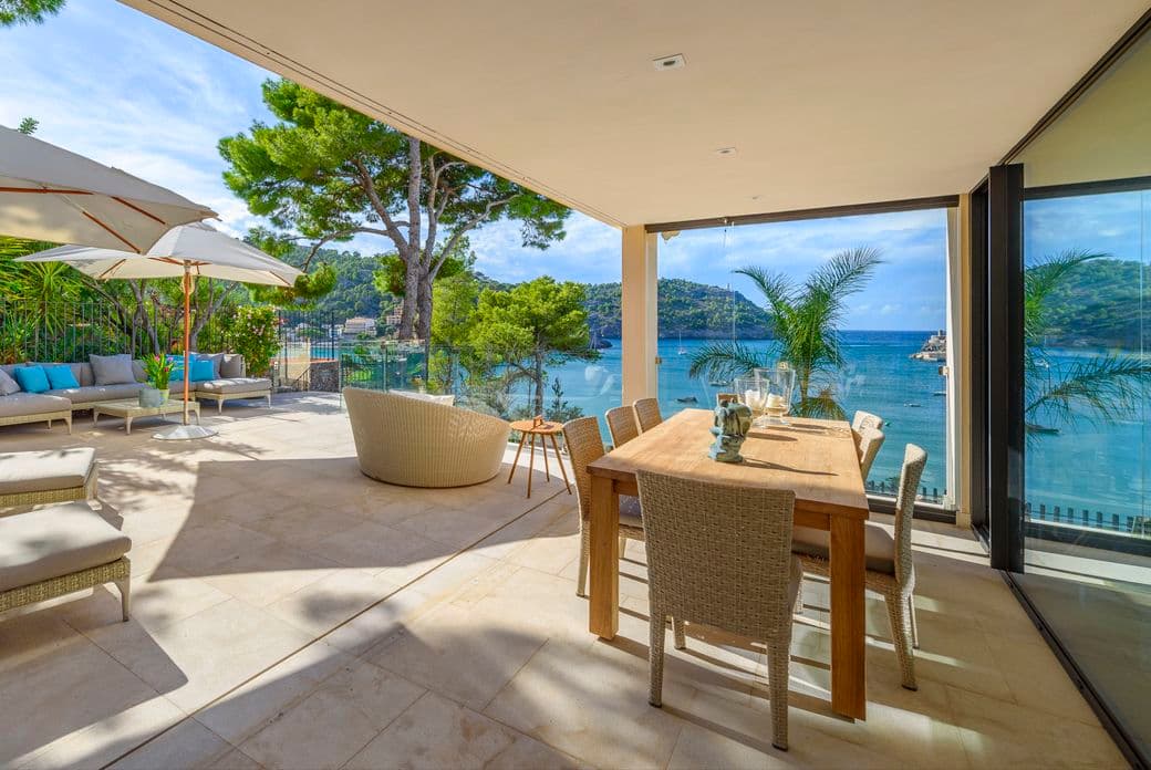 Scenic view from a hillside house overlooking the Port de Sóller bay in Mallorca, Spain, with the sparkling Mediterranean Sea in the background, sailboats in the harbor, and lush green hills surrounding the coastal village."