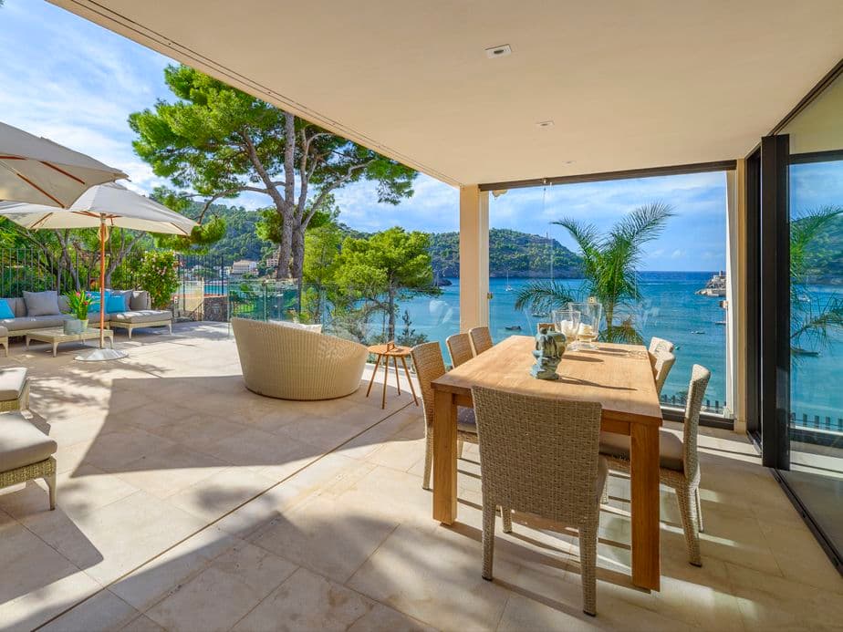 Scenic view from a hillside house overlooking the Port de Sóller bay in Mallorca, Spain, with the sparkling Mediterranean Sea in the background, sailboats in the harbor, and lush green hills surrounding the coastal village."
