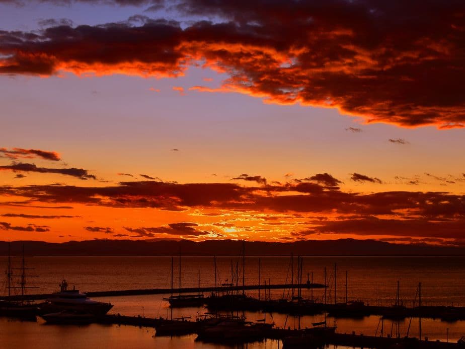 A vibrant sunset over a marina with silhouetted boats and a glowing orange and red sky, reflecting on the calm water.