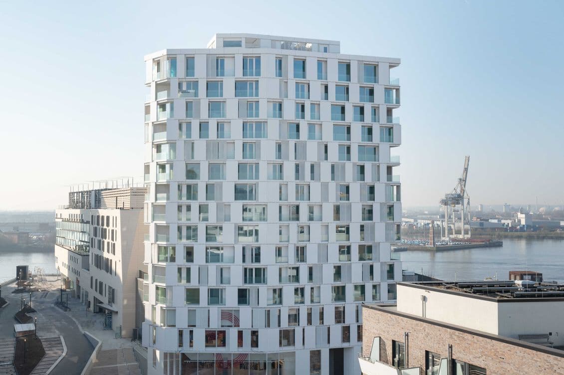 Modern white high-rise building with numerous windows, situated near a river with an industrial crane in the background. Clear blue sky.