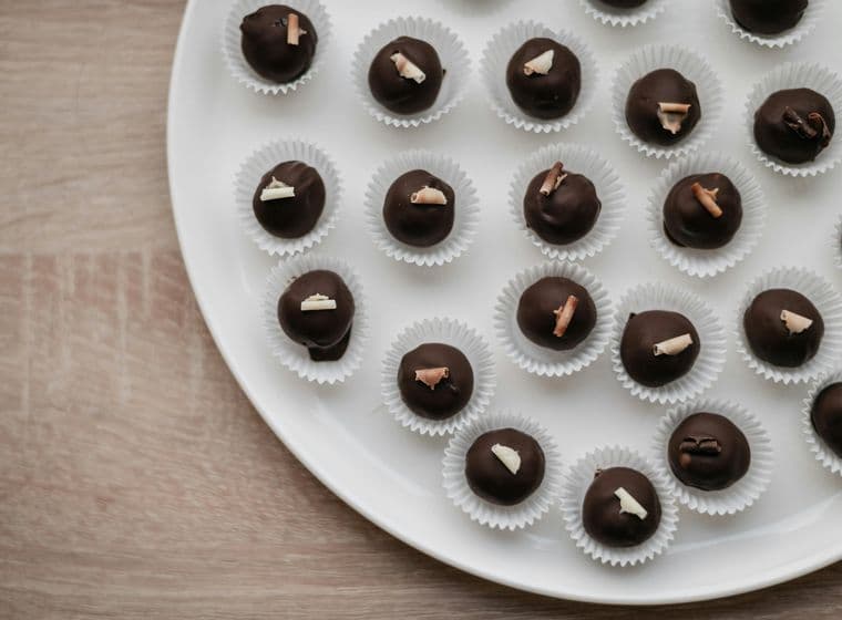 Des truffes au chocolat sur une assiette blanche. Chaque truffe est dans un moule en papier blanc et surmontée d'une spirale de chocolat. L'assiette est posée sur une surface en bois marron clair.