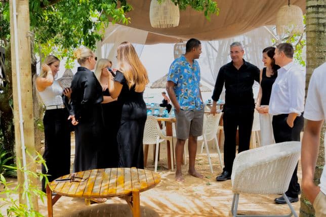 A group of people in casual and semi-formal attire converse at an outdoor gathering with a beach setting, surrounded by trees and wicker furniture.