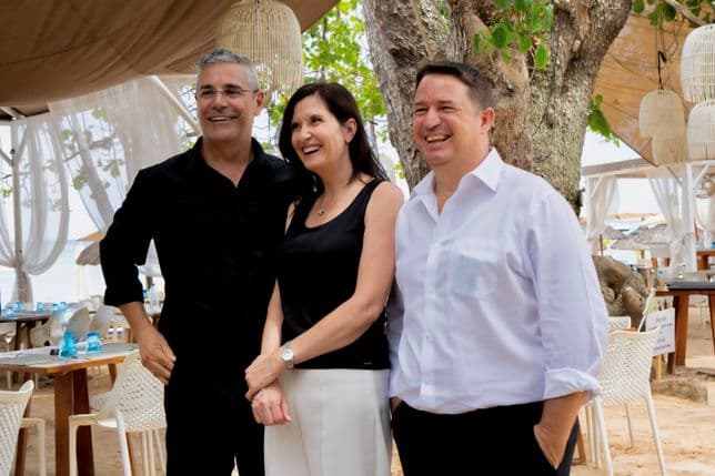 Three people smiling, standing together outdoors. They are dressed in black and white attire, with a beachside setting in the background.