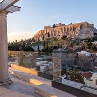 A terrace with columns and glass railings overlooks Athens, featuring the iconic Acropolis and Parthenon atop a rocky hill at sunset. Houses and trees fill the foreground.