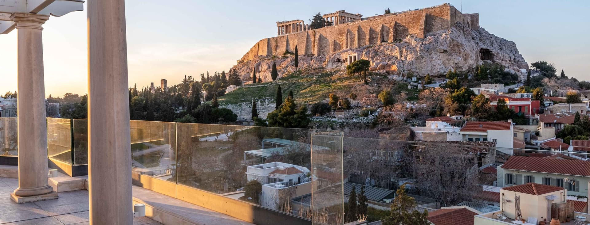 A terrace with columns and glass railings overlooks Athens, featuring the iconic Acropolis and Parthenon atop a rocky hill at sunset. Houses and trees fill the foreground.