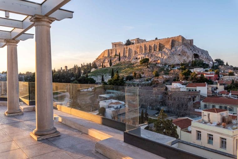 A terrace with columns and glass railings overlooks Athens, featuring the iconic Acropolis and Parthenon atop a rocky hill at sunset. Houses and trees fill the foreground.