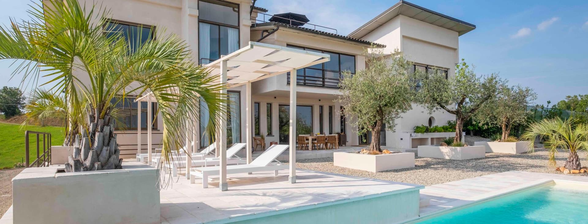 Villa moderna con ampie finestre, piscina e lettini prendisole. Circondata da palme e vegetazione, sotto un cielo azzurro e limpido.