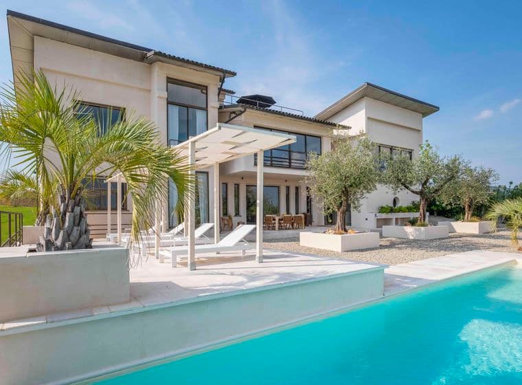 Villa moderna con ampie finestre, piscina e lettini prendisole. Circondata da palme e vegetazione, sotto un cielo azzurro e limpido.