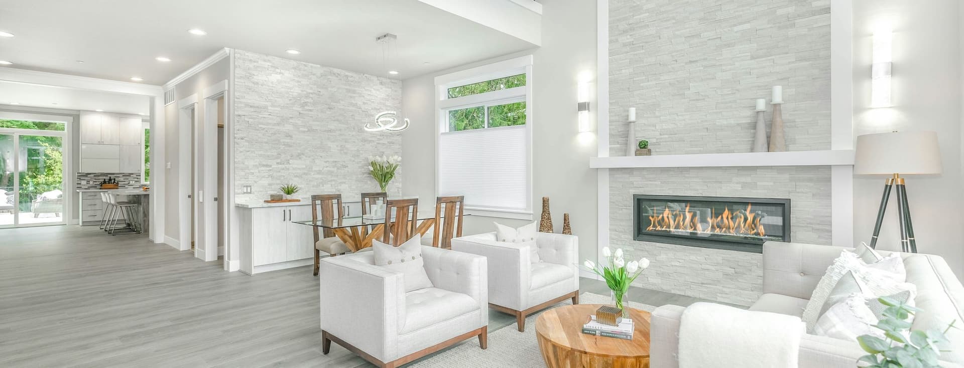 Bright, open-concept living space with white chairs, a fireplace, and a dining area. Light gray wood floors and stone accent walls.