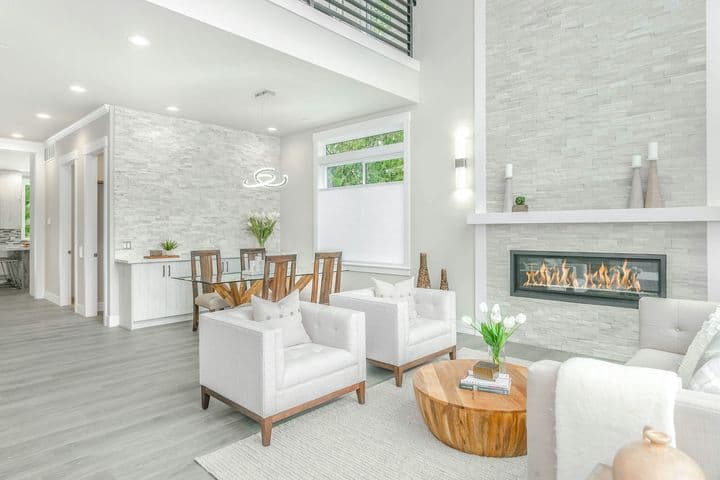 Bright, open-concept living space with white chairs, a fireplace, and a dining area. Light gray wood floors and stone accent walls.
