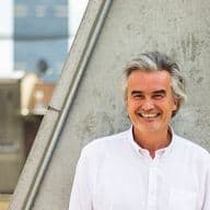Smiling man with gray hair in a white shirt stands against a concrete wall with buildings visible in the background.