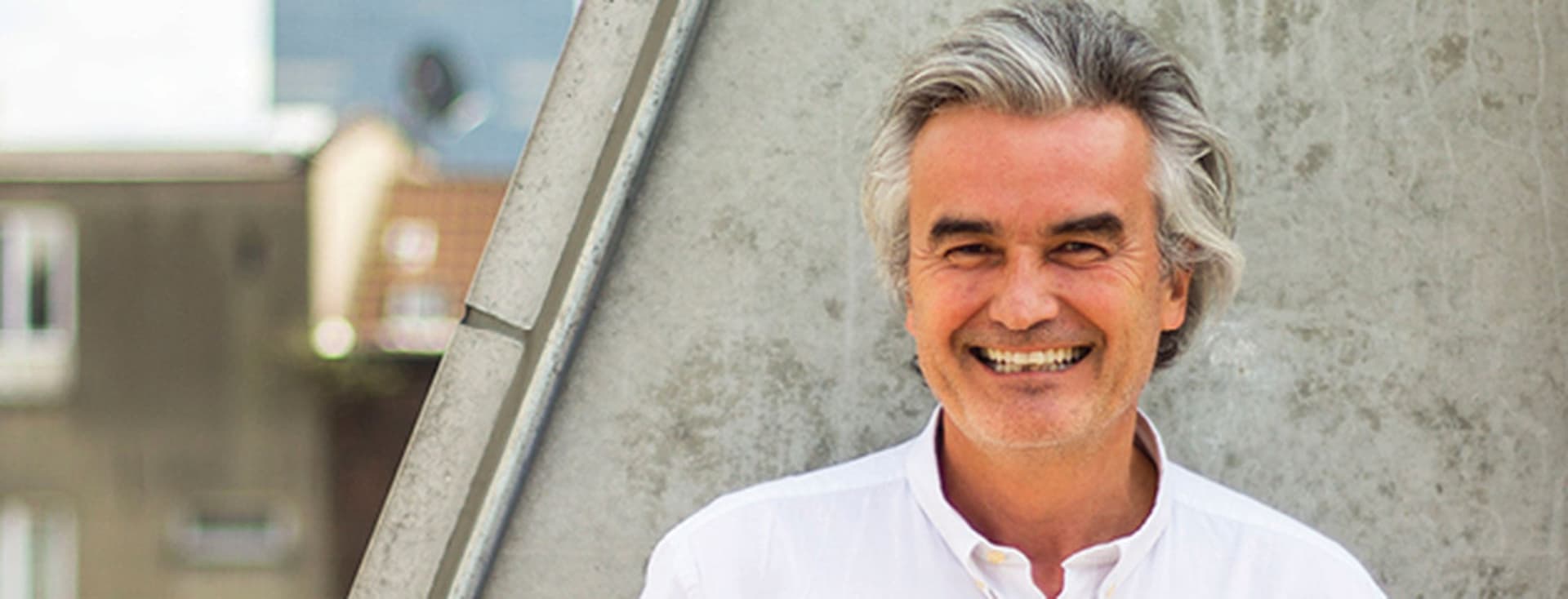 Un homme souriant aux cheveux gris, vêtu d'une chemise blanche, se tient devant un mur de béton avec des bâtiments visibles en arrière-plan.