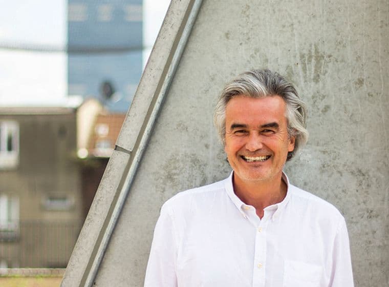 Un homme souriant aux cheveux gris, vêtu d'une chemise blanche, se tient devant un mur de béton avec des bâtiments visibles en arrière-plan.