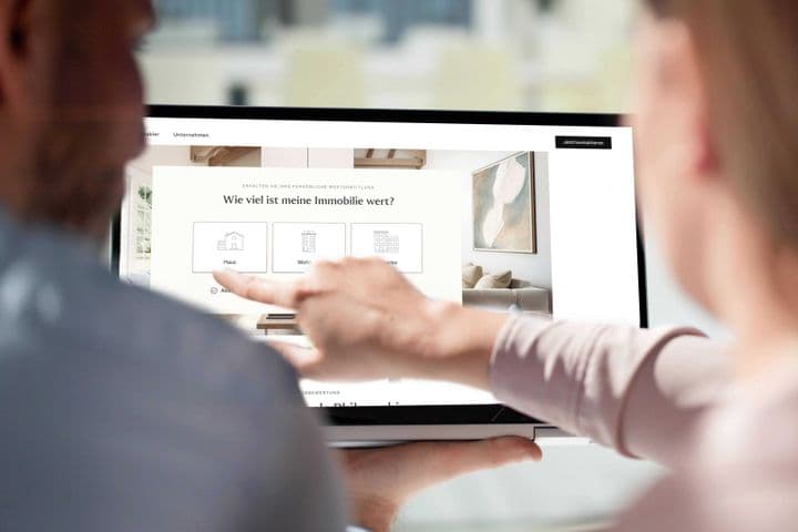 Two people pointing at a laptop showing a German real-estate valuation page reading "Wie viel ist meine Immobilie wert?"