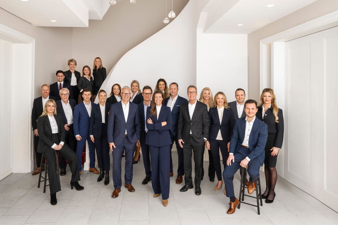 The Engel & Völkers Alstertal team poses professionally dressed in a modern, white room with a staircase in the background.