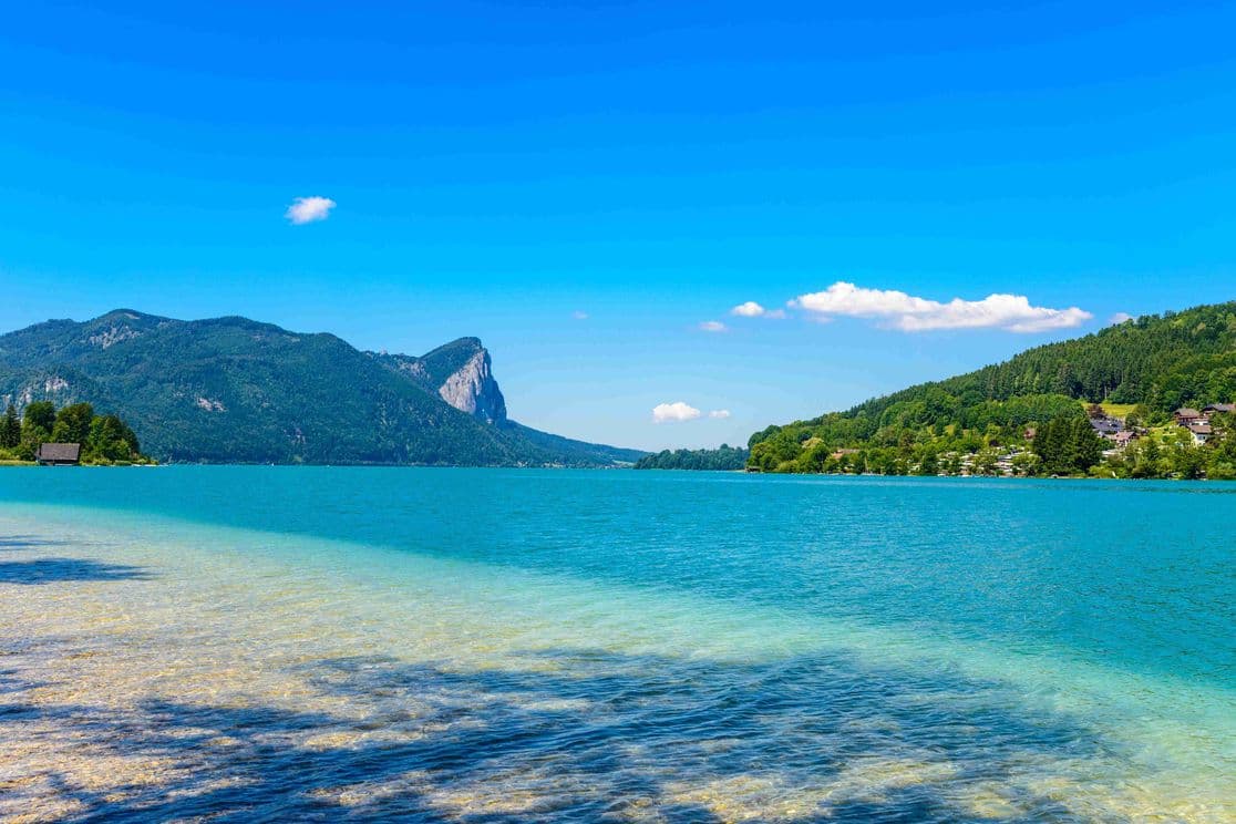Panoramaaufnahme des Mondsees mit türkisblauem Wasser, umgeben von bewaldeten Bergen und vereinzelten Häusern am Ufer unter blauem Sommerhimmel.