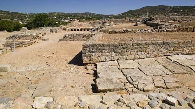 Ancient stone ruins with rectangular structures and pathways, set against a backdrop of hills and greenery under a clear sky.