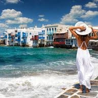 A woman in a white dress and hat looks out at the sea in Mykonos, Greece. White buildings with blue accents line the shore.